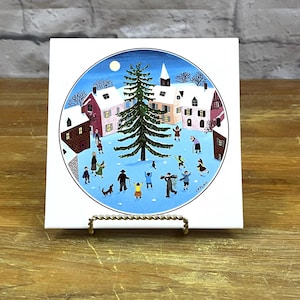 May include: A white ceramic tile with a blue and white circular design depicting a winter scene. The scene shows a small village with houses, a church, and a large evergreen tree. People are gathered around the tree, some are skating, and a dog is running. The text "LAPLAU" is visible in the bottom right corner of the tile.