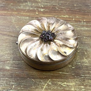 May include: A silver compact with a flower design and a purple rhinestone center.