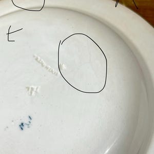 May include: A close-up of a white ceramic plate with a slightly curved rim. The underside of the plate shows embossed text and blue markings. The plate has a smooth, glossy finish and appears to be vintage.