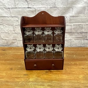 May include: A dark wood spice rack with eight clear glass jars with glass lids. The rack has two shelves and a small drawer. The rack is on a wooden surface with a white brick wall in the background.