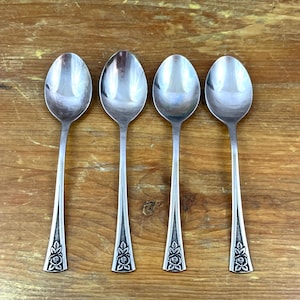 Könnte beinhalten: Vier silberne Löffel mit einem floralen Muster am Ende des Griffs liegen auf einer Holzoberfläche. Die Löffel haben ein klassisches, elegantes Design, ideal zum Servieren oder Essen. Die Löffel bestehen aus glänzendem, reflektierendem Metall.