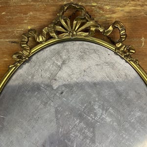 May include: A gold oval-shaped tray with a decorative ornate border. The border features a ribbon design with a bow at the top.