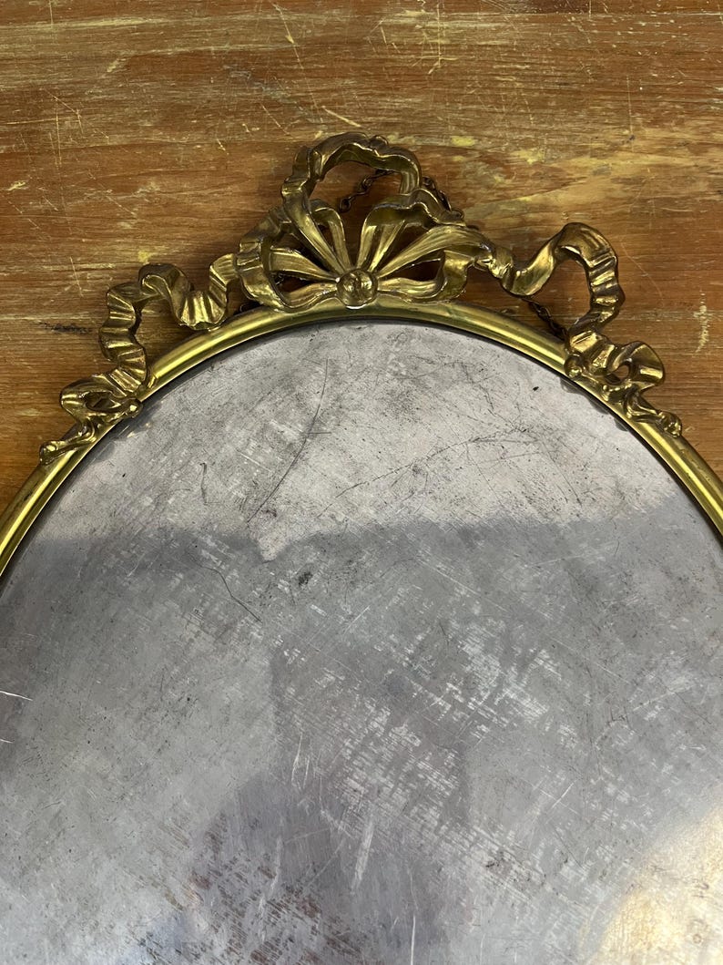 May include: A gold oval-shaped tray with a decorative ornate border. The border features a ribbon design with a bow at the top.