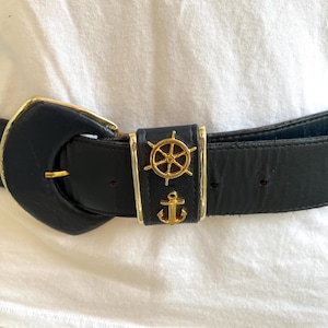 May include: A black leather belt with a gold buckle featuring a nautical theme. The buckle has a ship's wheel and an anchor design.