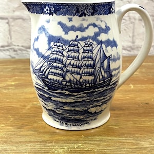 May include: A white ceramic pitcher featuring a blue transferware pattern of a sailing ship on the ocean. The text SS SHENANDOAH is printed near the base. The rim is decorated with a floral border, and the handle is curved on the right side.