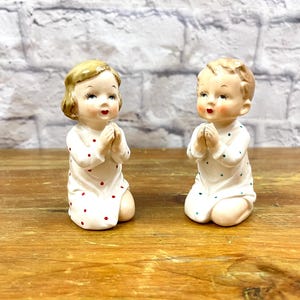 May include: Two vintage ceramic figurines of children kneeling in prayer. The figures are dressed in white robes with red and green polka dots. They have blonde hair and are positioned on a wooden surface.