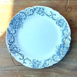 May include: A white ceramic plate with a scalloped edge and a blue floral design. The pattern includes flower clusters and scrolling vines around the rim. The plate is on a wooden surface.
