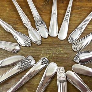 May include: An assortment of antique silver spoons and forks arranged on a wooden surface. The silverware features ornate floral and scroll designs, showcasing various patterns and styles. The spoons and forks are made of shiny, reflective metal.