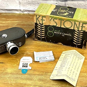 May include: A black Keystone 8 mm movie camera with a silver lens. The camera is in its original box, which is brown with green and white leaf designs. The box has the text "Keystone 8 MM Movie Camera" printed on it.