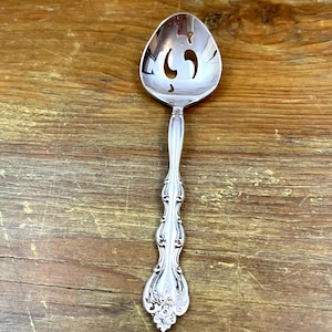 Può includere: Un cucchiaio da portata argentato con manico decorativo e coppetta forata. Il cucchiaio presenta un design ornato sul manico e un motivo floreale alla base. La coppetta ha tre fessure curve.