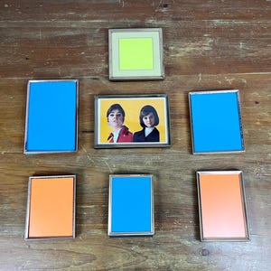 May include: A collection of vintage picture frames with silver-colored borders. The frames contain various colored backgrounds, including blue, orange, and a lime green. One frame holds a vintage photograph of two women. The frames are arranged on a wooden surface.