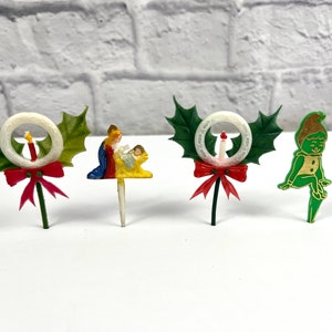 Peut inclure: Quatre décorations de gâteau de Noël vintage. L'une représente une couronne de houx avec un nœud rouge et une bougie blanche. Une autre représente une couronne de houx avec un nœud rouge et une bougie blanche. La troisième est une scène de la Nativité avec Marie, Joseph et l'enfant Jésus. La dernière est un lutin vert avec une ceinture dorée et un chapeau rouge.