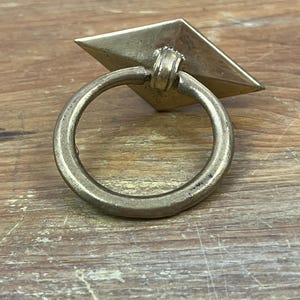 May include: A brass-toned metal drawer pull featuring a diamond-shaped backplate and a circular ring handle. The hardware has a weathered, metallic finish with visible signs of age and oxidation, showcasing a simple, functional design.