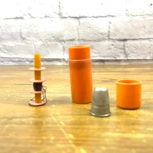 May include: An assortment of sewing notions on a wooden surface. The items include an orange container, a thimble, an orange cap, and a spool of thread. The background features a white brick wall.