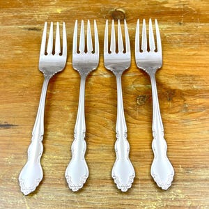 May include: Four silver forks with ornate handles are arranged on a wooden surface. The forks have tines and decorative detailing on the handles. The forks are made of a shiny, reflective metal.