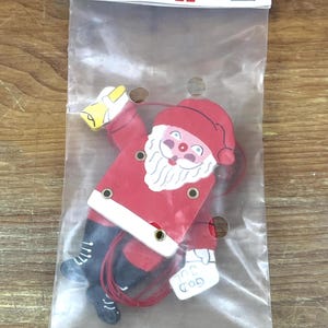 May include: A flat wooden Santa Claus ornament in a clear plastic bag with a header card labeled Larsen Danmark A/S. The Santa figure wears a red coat and hat, black boots, and holds a yellow bell and a white sign with the text GOD JUL.