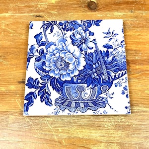 May include: Square ceramic tile with a blue and white floral pattern. The design shows a basket of flowers, with detailed elements and a traditional style. The tile is placed on a wooden surface.