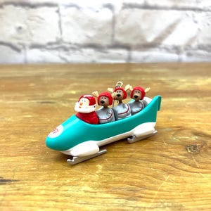 Puede incluir: Un adorno navideño con Papá Noel y tres renos en un trineo bobsleigh turquesa. Las figuras llevan cascos rojos y uniformes grises. El trineo tiene patines blancos y está sobre una superficie de madera.