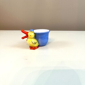May include: A blue and yellow ceramic egg cup shaped like a duck with an open beak. The cup is designed to hold an egg.