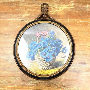 Op de afbeelding: Ronde lijst met een afbeelding van een mand met blauwe bloemen en een roze strik. De lijst is zwart met een gouden binnenrand en een decoratief bovenstuk. Het kunstwerk wordt gepresenteerd op een houten oppervlak.