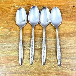 May include: Four silver spoons with oval bowls and ornate, floral-patterned handles are arranged on a wooden surface. The spoons are reflective, showing the surrounding environment. The spoons are likely for eating.