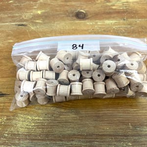 Puede incluir: Una bolsa de plástico transparente llena de 84 carretes de madera. Los carretes son todos del mismo tamaño y forma, y están dispuestos en un patrón aleatorio. La bolsa está etiquetada con el número "84".