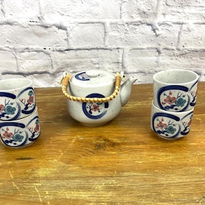 May include: A white teapot with a floral design and a brown wicker handle, along with four matching teacups stacked in pairs. The teapot and teacups have a white background with blue and pink floral designs.
