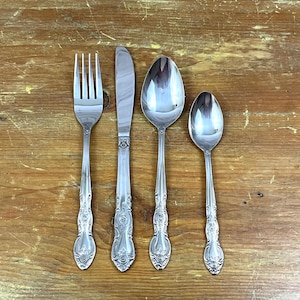 May include: A set of four silver-colored utensils: a fork, a knife, a large spoon, and a small spoon. The utensils have ornate, floral-like designs on the handles. The items are arranged on a wooden surface.