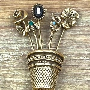 Puede incluir: Un recipiente dorado en forma de dedal contiene varios alfileres decorativos. Los alfileres presentan diseños florales y de insectos, incluyendo una rosa, una abeja y un camafeo. El dedal tiene una superficie texturizada, sobre fondo de madera.