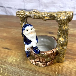 May include: A ceramic figurine of a gnome sitting on a well with a brown, gold, and blue color scheme. The well has a small pot on top.
