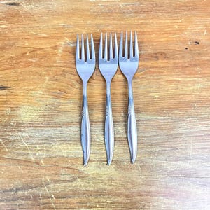 May include: Three silver forks with ornate designs on the handles, arranged in a row on a wooden surface.