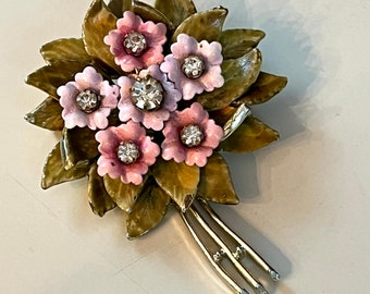 Puede incluir: Un broche vintage con un grupo de flores rosas con centros de pedrería, sobre un fondo de hojas verdes. El broche tiene un tallo dorado con tres puntas.