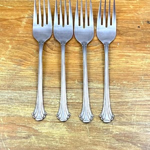 May include: Four silver forks with tines and decorative handles are arranged on a wooden surface. The forks have a classic design with a detailed pattern at the base of the handle. The background is a light brown, textured wood.