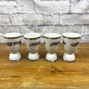 May include: Four white ceramic egg cups with a floral design in red, pink, and green. The egg cups have a rounded base and a decorative band at the top and bottom. They are arranged on a wooden surface.