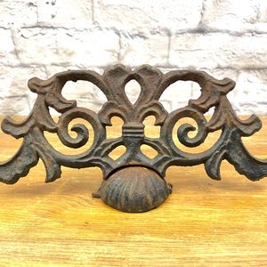 May include: Ornate, cast iron architectural salvage piece. The dark brown metalwork features a symmetrical design with scrolling details and a central finial. The piece has a semi-circular base and is likely a decorative element.
