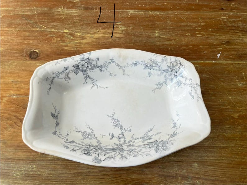May include: White rectangular serving dish with a floral design in grey. The dish has a slightly scalloped edge and is set against a wooden surface. The number "4" is written in black marker in the upper left corner.