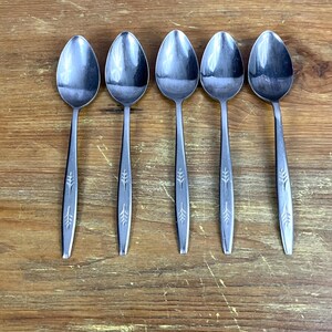 May include: Five silver spoons with a wheat design on the handle. The spoons are arranged in a row on a wooden surface.