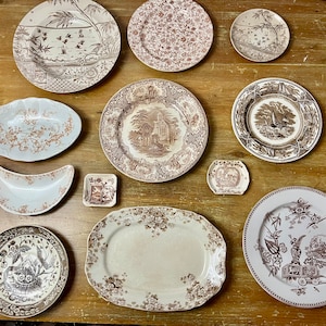 May include: A collection of 11 brown and white ceramic plates with various floral and geometric designs. The plates are arranged on a wooden surface.