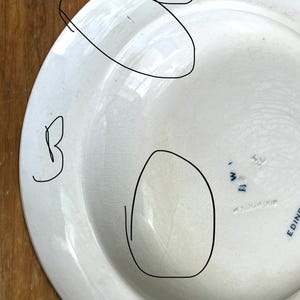 May include: A white ceramic plate with a slightly off-white tone. The plate shows a crack and some brown marks. The back of the plate features blue text, including "EDINBURGH" and "WEDGWOOD".