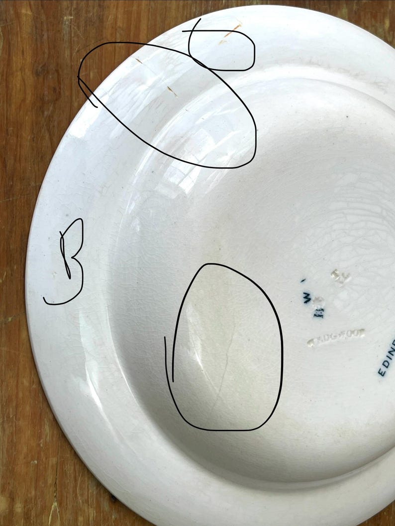 May include: A white ceramic plate with a slightly off-white tone. The plate shows a crack and some brown marks. The back of the plate features blue text, including "EDINBURGH" and "WEDGWOOD".