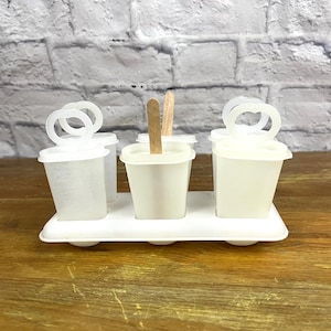 Peut inclure: Un moule à popsicles en plastique blanc avec quatre compartiments individuels et des bâtonnets en bois. Le moule a une base blanche et est conçu pour contenir des popsicles.