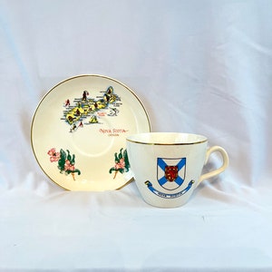 May include: A white teacup and saucer with a gold rim. The saucer features a map of Nova Scotia, Canada, with various landmarks and activities depicted. The teacup has the Nova Scotia coat of arms with the text "Nova Scotia" underneath.