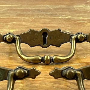 May include: A brass drawer pull featuring a central keyhole escutcheon plate with a decorative bow-shaped silhouette and a curved drop handle. The metal hardware has a tarnished finish with visible patina, showcasing a classic design with pointed ends.