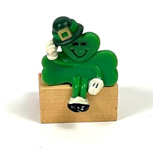 May include: A green shamrock figure with a smiling face, wearing a green hat with a gold buckle, and white gloves. The figure is sitting on a small wooden box. A festive St. Patrick's Day decoration.