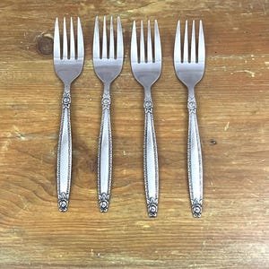 May include: Four silver forks with ornate floral and geometric designs on the handles are arranged on a wooden surface. The forks have four tines each and a decorative pattern near the base of the tines.