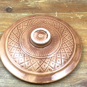 Könnte beinhalten: Brauner Keramikdeckel mit kreisförmigem Design und zentralem Knopf. Der Deckel hat ein geprägtes Muster aus Rauten und Linien und ist wahrscheinlich für einen Topf oder eine Auflaufform. Der Knopf ist cremefarben.