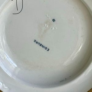May include: A white ceramic plate with a raised rim. The underside displays the word "EDINBURGH" in blue, alongside other marks. A large black "D" is hand-drawn on the plate's surface.