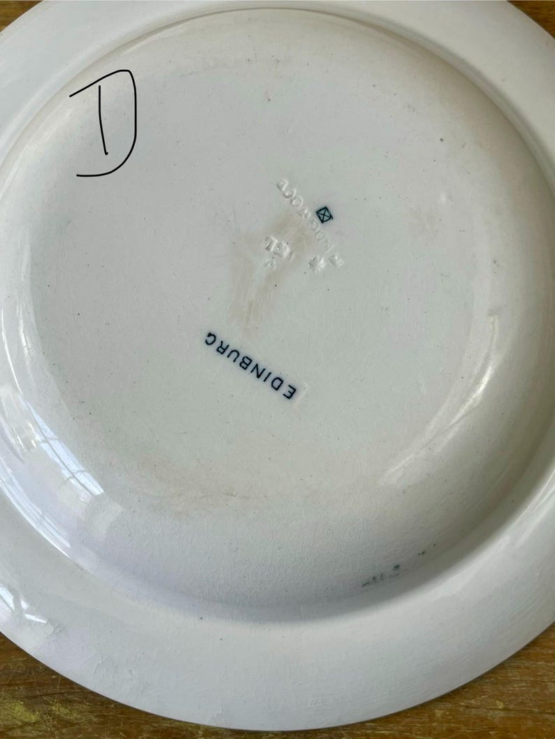 May include: A white ceramic plate with a raised rim. The underside displays the word "EDINBURGH" in blue, alongside other marks. A large black "D" is hand-drawn on the plate's surface.