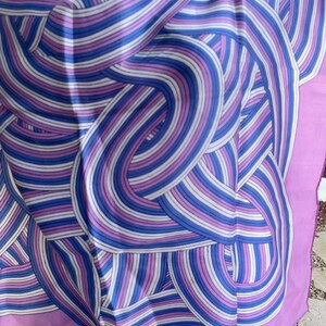 May include: A silk scarf with a repeating pattern of curved lines in shades of blue, purple, and white. The scarf has a solid lavender border. The design creates a sense of movement and visual interest.