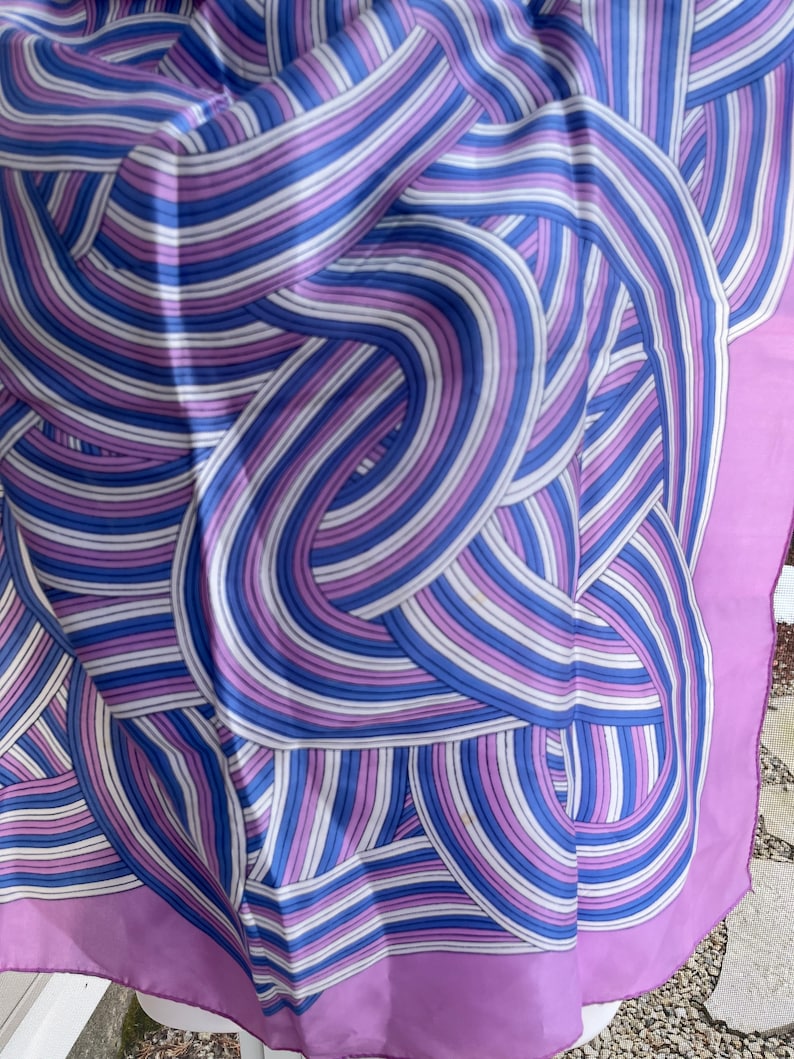 May include: A silk scarf with a repeating pattern of curved lines in shades of blue, purple, and white. The scarf has a solid lavender border. The design creates a sense of movement and visual interest.
